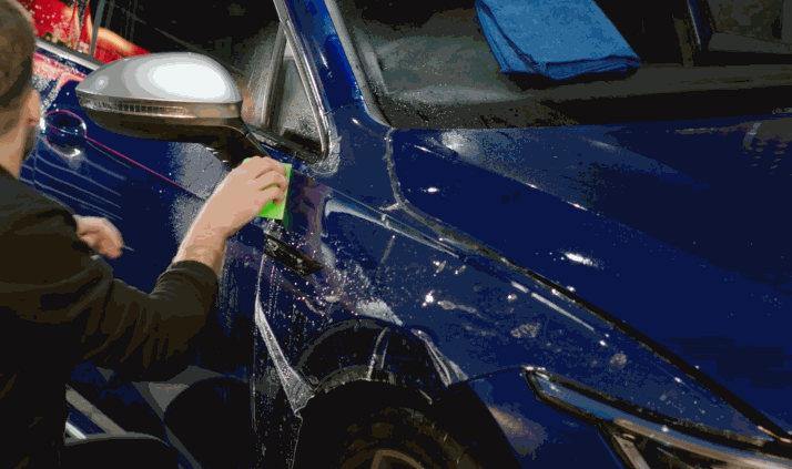 Installer uses a green squeegee to apply paint protection film to a blue car door with a soapy solution for precise positioning. The process removes air and moisture to create a clear protective layer.
