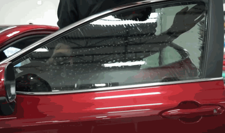 Red car door with partially installed window tint showing small water droplets trapped between the glass and tint film during application. The technician stands above the door working inside the vehicle while the window tint settles evenly across the side window. The workshop lighting reflects across the glass surface.