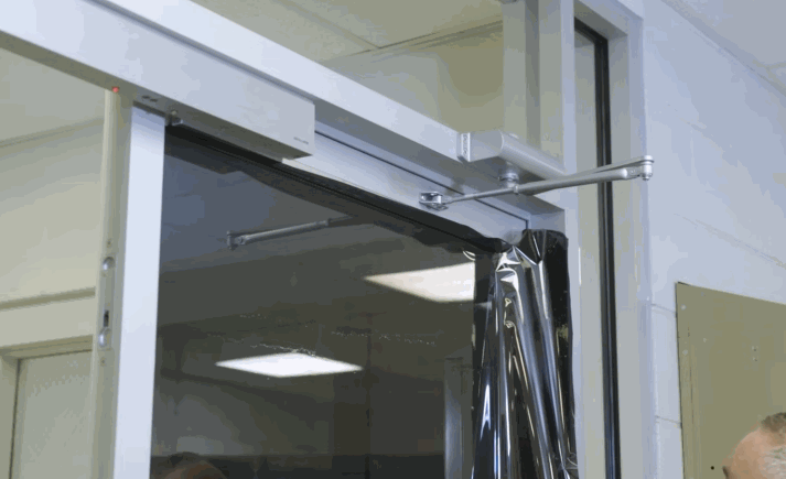 Close up of a glass door as dark commercial window tint film is carefully installed near the top hinge and closing mechanism. The reflective surface shows overhead lights while the film is being aligned.