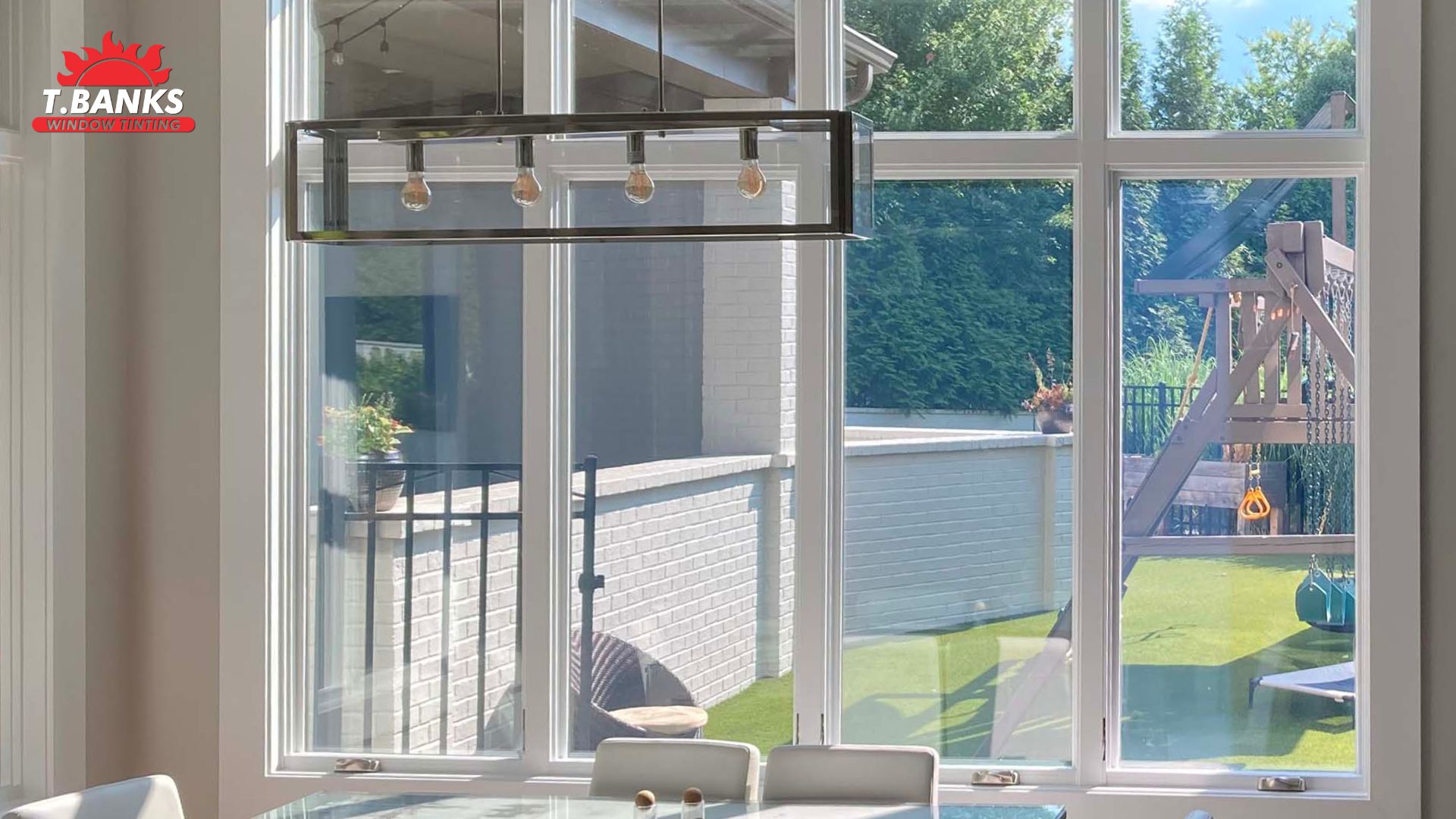 Bright dining area with large windows overlooking a backyard playset and patio, featuring a subtle tint applied for residential window tint to reduce glare and heat. A modern rectangular light fixture hangs above a table with chairs. The tinted glass keeps the space روشن and comfortable while filtering sunlight.