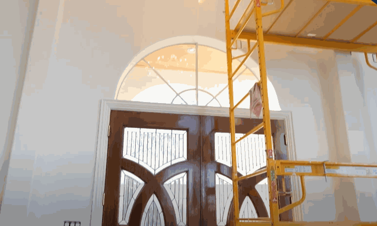 Interior entryway with tall arched window above dark wooden double doors while a yellow scaffold stands to the right during installation of residential window tint. Natural light filters through the untreated glass highlighting the height and shape of the window. This shows a work in progress before tint is applied.