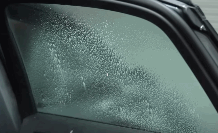 Close up of a car window covered in fine water droplets as window tint film settles and adheres to the glass surface. The texture highlights the preparation stage of window tint application before final smoothing.