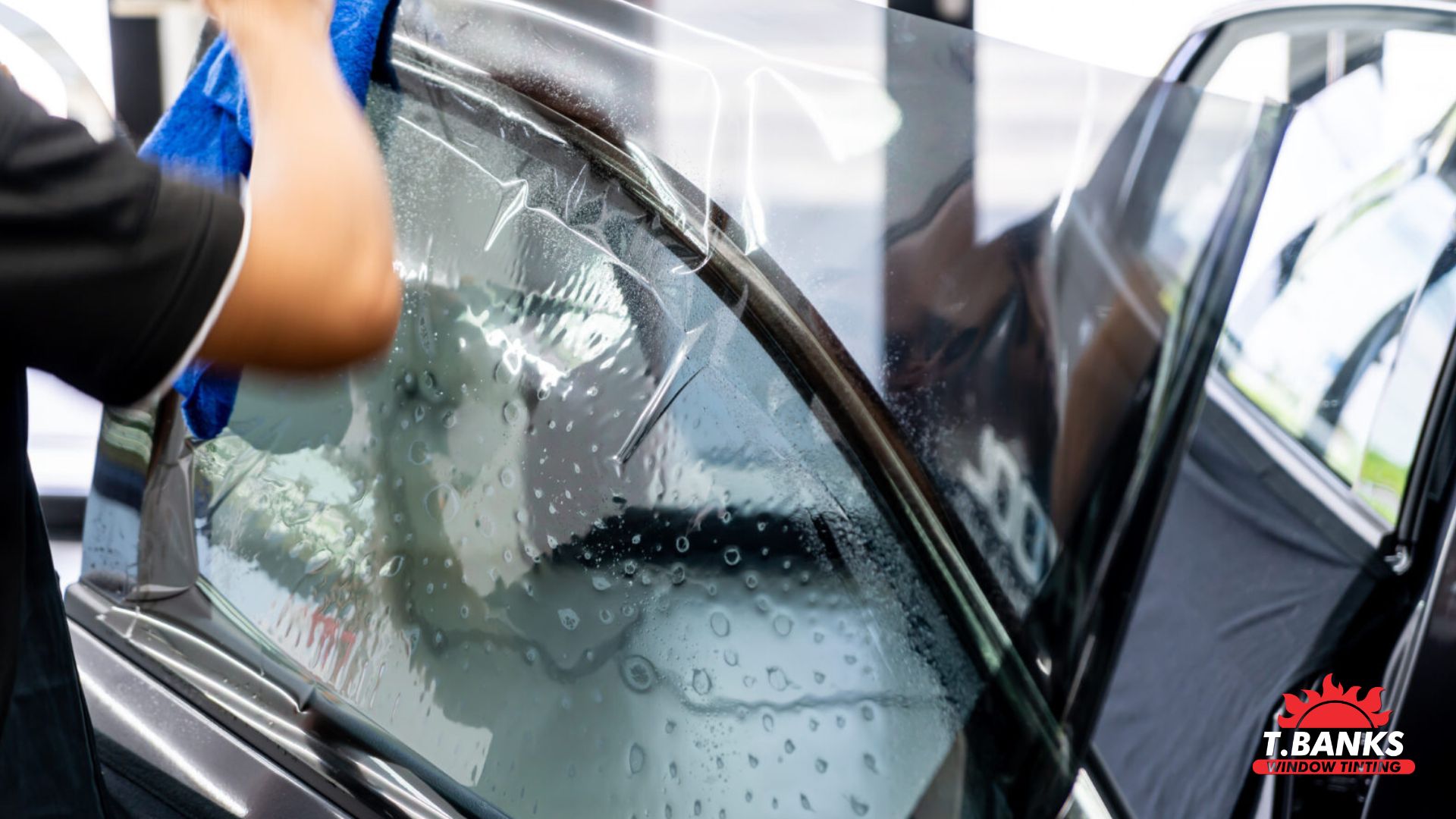 Technician applies window tint film to a car door glass covered in water droplets using a blue cloth to smooth it into place. The process shows window tint installation in progress with the car door open and film partially adhered.