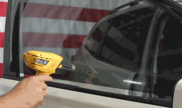 Hand uses a yellow heat gun to shrink and fit ceramic window tint onto a car side window, smoothing the film as it conforms to the glass. The dark tint gradually tightens against the surface for a seamless finish.