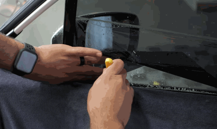 Close up of hands trimming ceramic window tint along the edge of a car window using a small blade while moisture beads on the glass. The film is carefully aligned for a precise and clean installation.