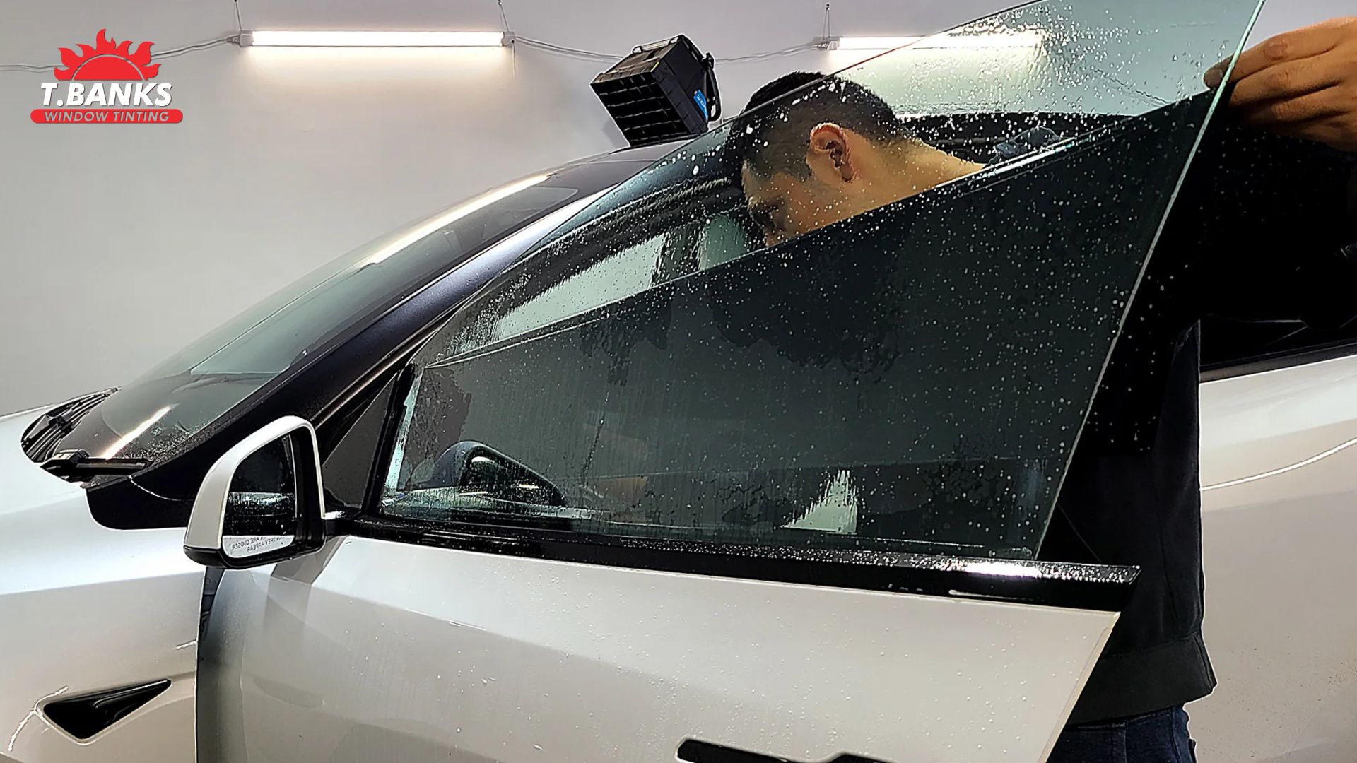 Technician installs a large sheet of ceramic window tint onto a car door window covered in water droplets inside a workshop.