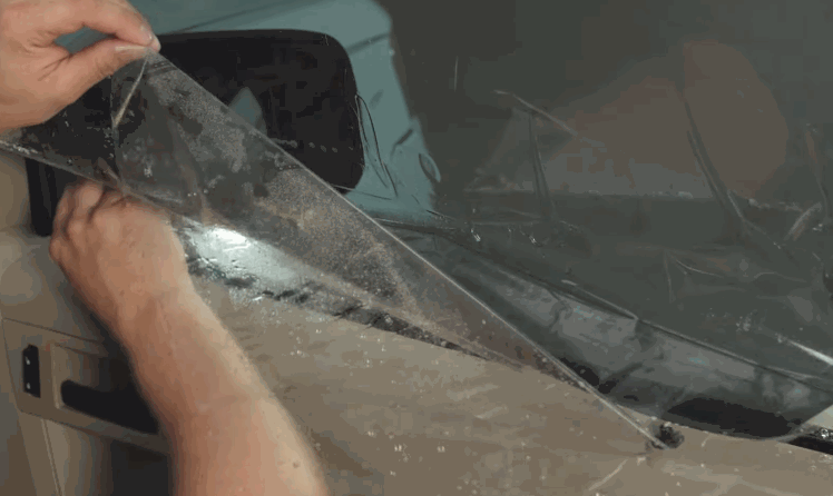 A technician peels and positions ceramic window tint on the side window of a beige vehicle. Water and film edges show the tint being prepared for installation.