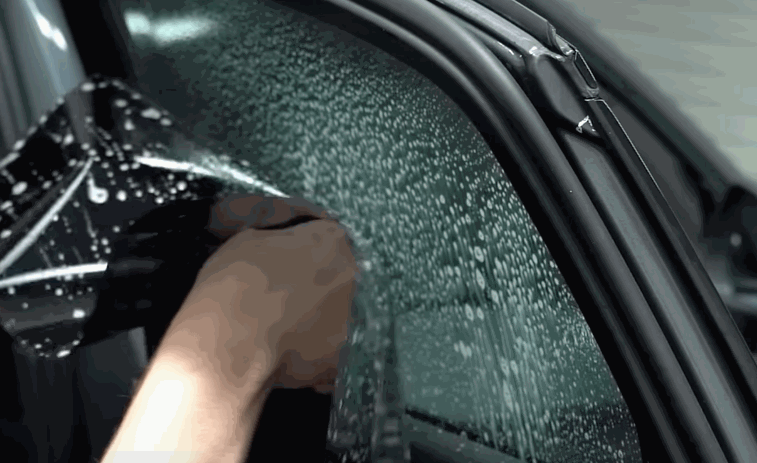 A technician installs window tint on a car door glass covered with water droplets. Hands guide the film into place along the curved edge of the window for a smooth fit.