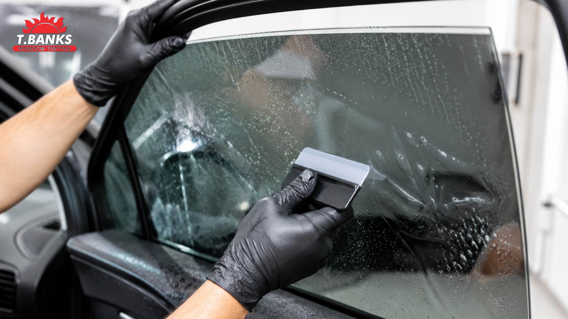 A technician in black gloves applies window tint to a car door window using a squeegee. The text "T.BANKS WINDOW TINTING" appears in the top left corner.