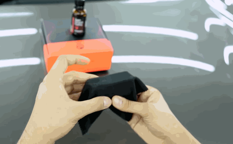 Hands prepare a black applicator pad near a ceramic coating bottle on a glossy vehicle hood.