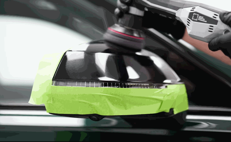 Detailer uses a machine polisher on a black side mirror cap while the surrounding edges are protected with green masking tape.