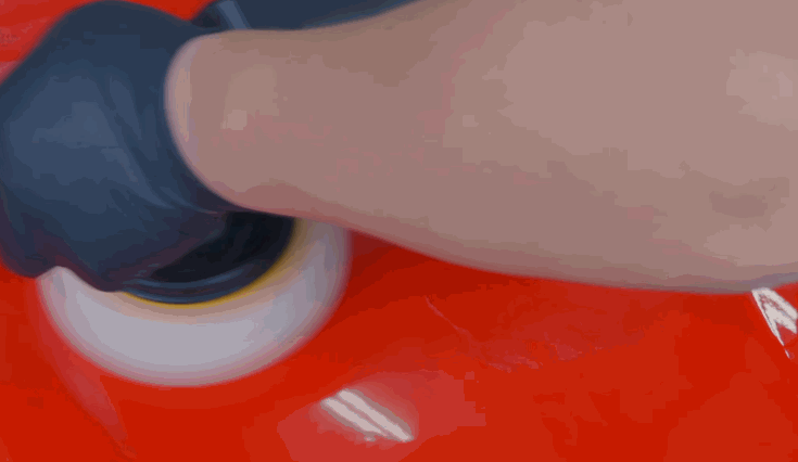 Close view of a machine polisher working across glossy red automotive paint. The polishing pad moves over the panel to refine the finish before protective treatments are applied.